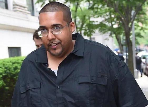 Un homme aux cheveux courts et aux lunettes se promène à l'extérieur, vêtu d'une chemise noire boutonnée. La verdure et un bâtiment sont visibles en arrière-plan. C’est Hector Monsegur, connu aussi sous le pseudo de Sabu. Hacker arrêté par le FBI, qui a coopéré, et mené à l’arrestation d’autres hackers