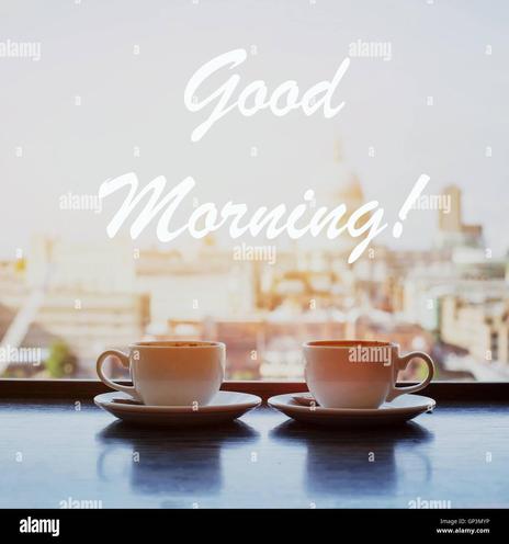 An image of two tan/peach colored cups and saucers atop a bluish table ... a Good Morning! message is conveyed, written in white coloration. Hazy background looks like a window's view outside (of the neighborhood), possibly.