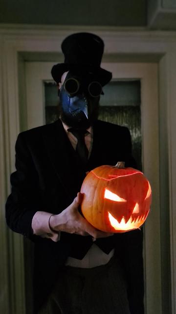 Spooky gent in top hat, beaked plague doctor mask, black jacket, pink shirt, black tie holding out a lit pumpkin