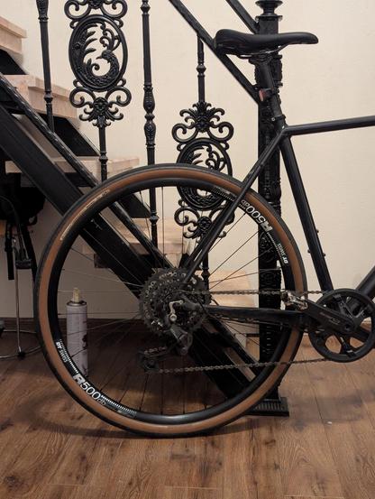 Велосипед, стоящий у лестницы с красивыми коваными перилами. Колесо велосипеда с коричневой покрышкой и чёрной рамой выделяется на фоне деревянного пола.