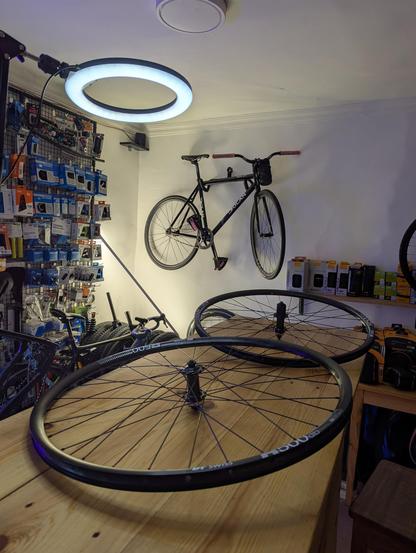Мастерская по ремонту велосипедов. На переднем плане лежат велосипедные колёса на деревянном столе, а на стене висит велосипед. В углу видны различные аксессуары и запчасти для велосипедов. Над столом висит кольцевая лампа, освещающая рабочее пространство.