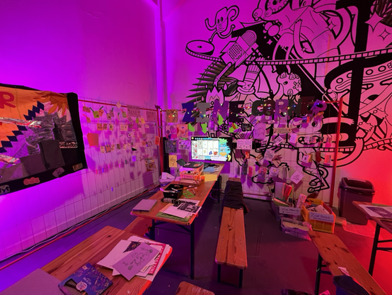 a photo showing a monitor hovering above a printer. EZM is on the monitor. the area around that is covered in odds and ends of art and zines. cut out letters that read “zine corner” are decorated with christmas’s lights. it is a mood!