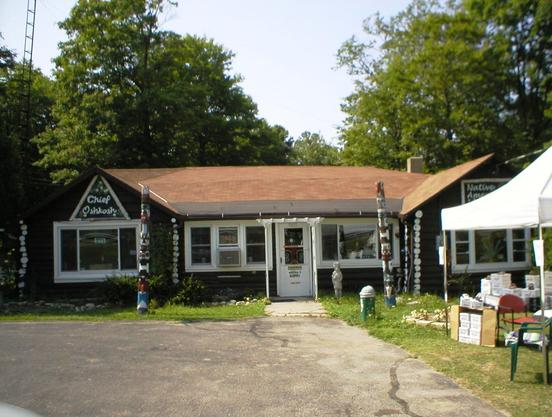 The image shows the Chief Oshkosh Trading Post, a rustic log cabin-style building with a brown roof, located on Highway 42 in Door County, Wisconsin. The building has a sign that reads 'Chief Oshkosh' on the left side and 'Native American' on the right side. There are totem poles and various Native American artefacts displayed outside. A white tent is set up on the right side of the building with boxes and items for sale. Lush green trees surround the area.