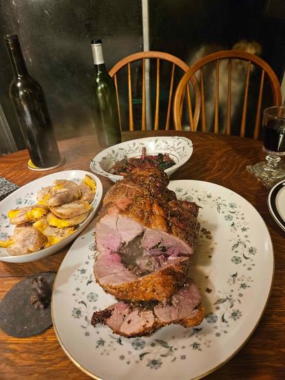 a holiday feast laid out on floral china platters: leg of lamb roast, sliced medium-rare; smashed gold potatoes; sauteed red chard; homemade blackberry wine. 