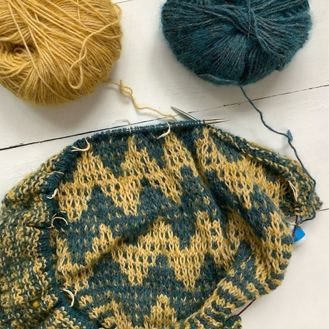Flat lay view of a triangle shawl with mosaic patterning on a circular knitting needle, together with yellow & petrol yarn on a white plank background