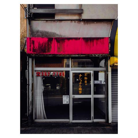 The photo shows a small, abandoned storefront, likely a former real estate office. The faded pink awning above is streaked with grime and wear, contrasting with the stark glass door below. The shop's interior is completely empty, with curtains pulled aside to reveal bare floors and walls. Yellowed lettering on the door and windows advertise services that are no longer offered. Reflections of the street outside fill the vacant space, amplifying the sense of abandonment.