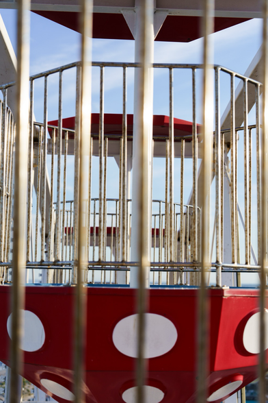 A view of empty Ferris wheel cars shot from another Ferris wheel car. Strong white vertical bars cut through the foreground of the image with bits of the cars red and white polka dots sticking through form the bottom and background. 