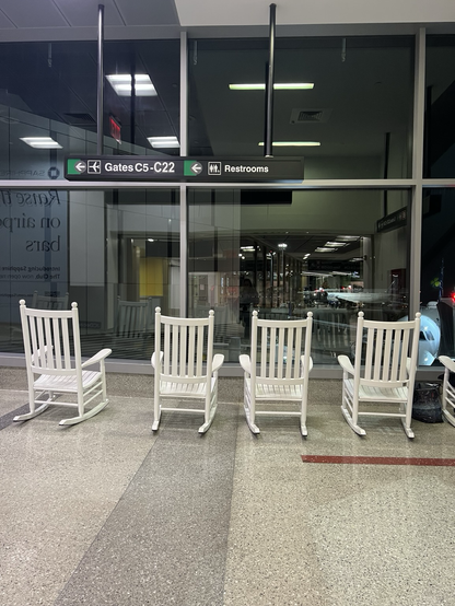 A row of white painted wooden rocking chairs, in between terminals at Boston airport. They look out on the aircraft …. Very nice!!