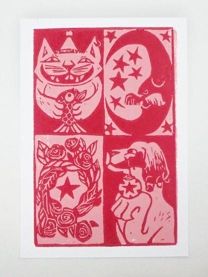 A hand printed lino cut card, the recto side. It is printed in two colours: Red and pink. You see a grid of four parts. From the top left to the bottom right: A cat with a hat holding a fish, a moon with a moustache, a Christmas wreath and a dog with a bone in its mouth from which hangs a Christmas bauble. 