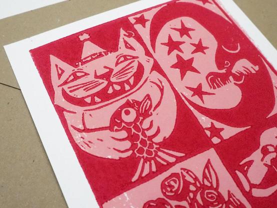 Detail of the hand printed lino cut card, the recto side. It is printed in two colours: Red and pink. The detail is focused on the cat with a hat holding a fish and the moon with a moustache.