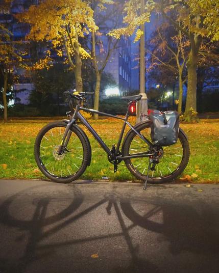 Ein Fahrrad steht am Rand eines breiten Radwegs. Der Blick ist auf die Linke Fahrradseite gerichtet. Im Vordergrund ist Asphalt, dahinter eine Wiese mit Bäumen. Die Szene ist von einer Laterne orange beleuchtet.
Das Fahrrad ist grau mit grauen Satteltaschen, es leuchten 2 rote Rücklichter. Das Rad wirft einen Schatten in den Vordergrund. 