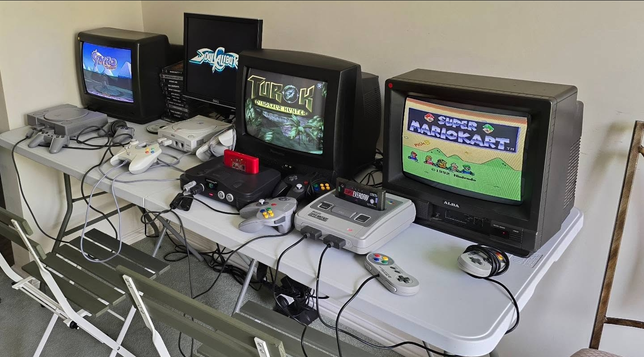This image shows a retro gaming setup featuring several classic gaming consoles and CRT televisions. Here's what's visible:

1. PlayStation 1 (PS1): The first console on the left, with two grey controllers connected.

2. Sega Dreamcast: The second console, identifiable by its white colour and the distinctive controller design.
L

3. Nintendo 64 (N64): The third console with a black body and grey controllers. It has a red expansion pack on top.

4. Super Nintendo Entertainment System (SNES): The fourth console, featuring two SNES controllers, and a "Super Mario Kart" game cartridge inserted.

Each console is connected to a CRT television or monitor displaying a relevant game screen. The overall setup evokes a sense of nostalgia, aimed at recreating the retro gaming experience.