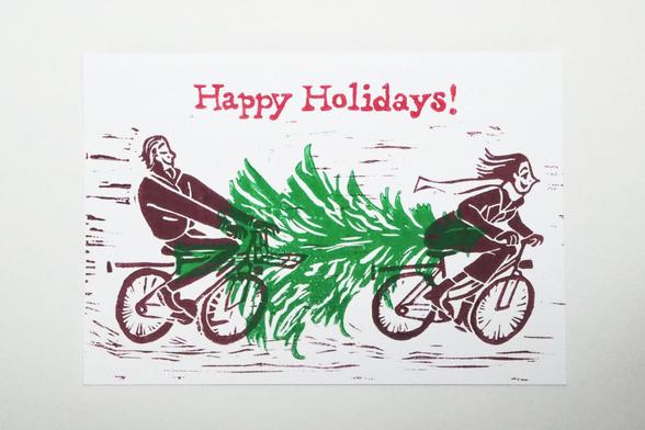 A hand printed lino cut holiday card, with a man and a woman who are riding their bicycles, printed in a dark maroon red colour. In between them is a bit of space and there lies a Christmas tree, printed in green. They are transporting the tree together. Above them, it reads “Happy Holidays!” printed in red. 