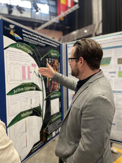 Tobias Hank presenting their poster on using spectroscopy to measure nitrogen content in crop canopies. He is looking and gesturing at the poster with his right hand. The poster shows some text and diagrams along the leafs of a plant.