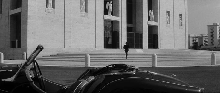 a stylish dark car parked in front of a large museum gallery building, the film is black and white