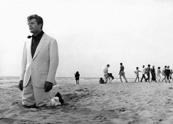 a man in a white suit on his knees, looking exhausted, on the beach, people hanging out in the sand behind  him