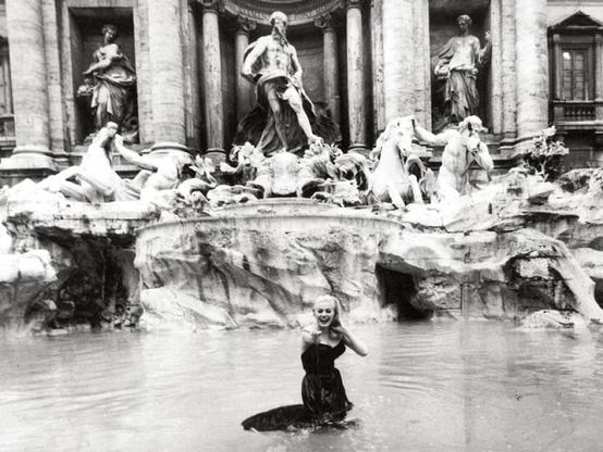 the blonde woman laughing, she isup to her knees in the water in a fancy  fountain with statues behind her, she is looking at the viewer , extending her hand
