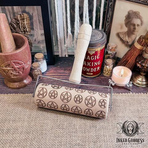 A small rolling pin sits on a burlap-covered surface. The Pentacle Rolling Pin has a handle like a paint roller, and it has pentacles of alternating sizes laser-engraved on the roller. This is an exclusive product from Inked Goddess Creations.