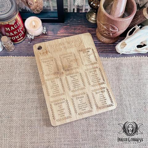 A small cutting board sits on a burlap-covered surface. The face of the Kitchen Witch Herb Magick Cutting Board is laser-engraved with various energies and a few of the herbs that embody them. It is made of bamboo and has a hole drilled near one corner for hanging. This is an exclusive product from Inked Goddess Creations.