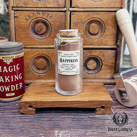 A clear glass vial of Kitchen Witch Magickal Culinary Blend: Happiness is displayed on a low wooden platform. The vial includes the ingredients list: sugar, cocoa powder, cinnamon, ginger, nutmeg, allspice, and orange peel. The vial is sealed with a cork plug, and it has a yellow cord around its neck with a silver peace heart charm attached. This is an exclusive product from Inked Goddess Creations.