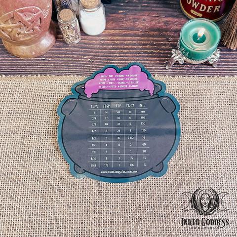A round refrigerator magnet sits on a burlap-covered surface. The face of the Cauldron Magnetic Kitchen Conversion Chart is a round dark grey cauldron with purple froth flowing out of it. It's printed with various measurement conversions. This is an exclusive product from Inked Goddess Creations.