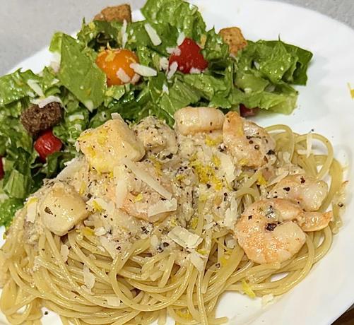 Angled closeup of a pile of spaghetti on a white plate, topped with scallops and shrimp in a bit of sauce. Green salad behind topped with multicoloured cherry tomatoes and croutons. All garnished with grated Parmesan,pepper and lemon zest. 