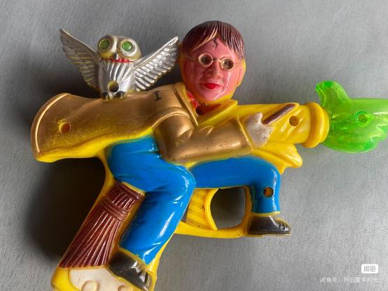 A chinafake toy gun that resembles Harry Potter on a broomstick with an owl. One of his hands is pointing at something, and is very large and made out of translucent green plastic with an LED inside. The whole thing is very off-looking, has a weird paint job, and Harry's face is that of an eldritch horror. The whole toy is covered in  minor damage from wear (scratches, paint worn down, etc) and small amounts of grime and dirt here and there, presumably from storage.