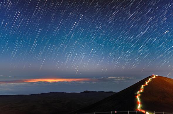Star trails streak across the sky above the summit of Maunakea in this image, which shows the surroundings of the Gemini North telescope, one half of the International Gemini Observatory, supported in part by the U.S. National Science Foundation and operated by NSF NOIRLab. The long exposure time of this photograph, coupled with the rotation of Earth, has caused the stars to streak into a multitude of graceful arcs. To the right of the image a much more earthly trail is visible; hikers with head torches have illuminated the final stretch of the Humuʻula Trail which winds up to the summit of Maunakea.

One of the streaks of light in the skies above Maunakea didn’t originate from a distant star, but rather from something much closer to home. A passing airplane has left a dashed trail at the lower left of this image, pointing the way to the glowing patch of cloud illuminated by the city lights of Hilo, Hawai‘i.