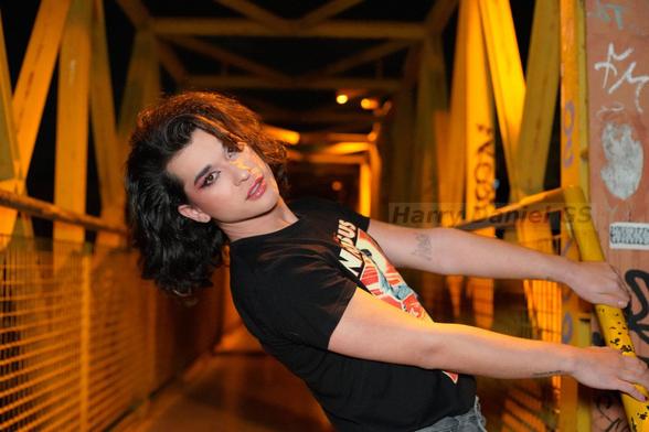 Modelo sujetada con ambas manos a una barandilla amarilla en un puente amarillo, inclinada hacia detrás.
Lleva una camiseta de Ninjisus (Jesús ninja)
Pelo suelto negro que le tapa algo la cara con labios y ojos pintados de rosa. Eyeliner negro. Esfe noche. Le ilumina un foco.
Mira a la cámara.
