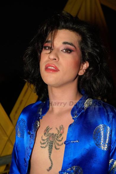Modelo con chaqueta azul potente con motivos asiáticos dorados abierta. Tatuaje de escorpión en el pecho.
Pelo suelto negro que le tapa algo la cara con labios y ojos pintados de rosa. Eyeliner negro. Está en un puente amarillo, de noche. Le ilumina un foco.
Mira a la cámara.