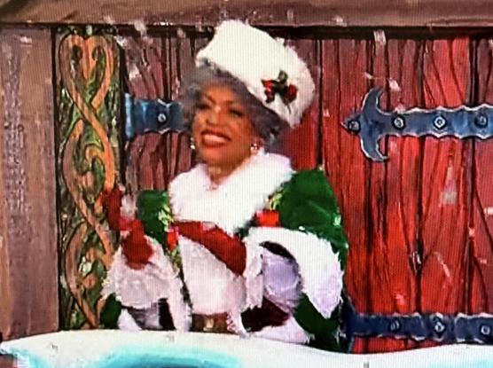 A smiling Ms Claus in a festive green and white outfit with red gloves and a hat, standing in front of a decorated background. Snow-like particles are falling around her, adding to the holiday atmosphere.