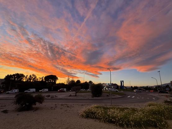 A scenic view of a sunset with vibrant orange and purple clouds, overlooking a roundabout with vehicles. Silhouettes of trees and shrubs are visible in the foreground.