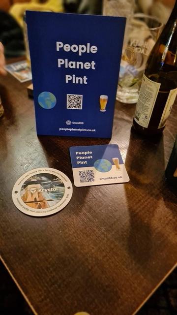 Picture of bar table with small standing blue sign for People Planet Pint, 2 coasters one for the aforementioned, one for Krystal web hoster, out of focus behind is pint glass, hefeweizen brown bottle and rest of table