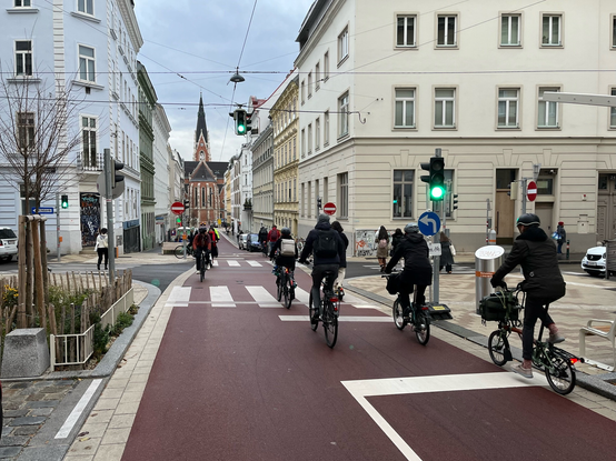 Fahrradstraße in Wien, im Hintergrund die Elisabethkirche, roter Asphalt, viele Radfahrende, grün leuchtende Ampel