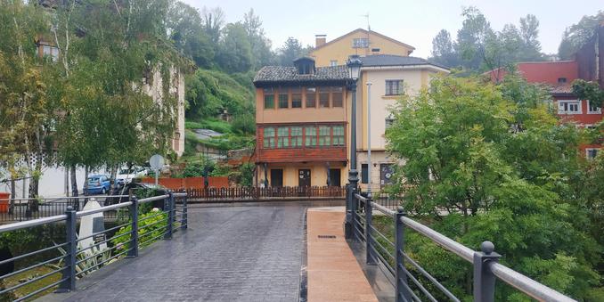 A horizontal coulour photo of a modern cobblestone footbridge over a river, wet after the rain. At the end of it and across the street it meets, there are two-storey houses woth windows on their façades. Behind the houses there are trees and emerald green vegetation.