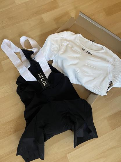 Foto einer schwarzen Rennradhose mit weißen Trägern, und eines T-Shirts aus einem Netzstoff in weiß.