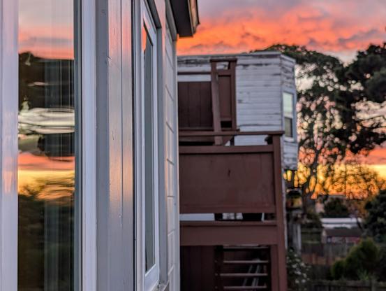 Photograph of a post-rainstorm sunrise with clouds painted in vivid coral, orange, and gold. The sunrise is reflected in a window, the view distorted by curvature in the window. CC BY-SA Kate Zimmerman.