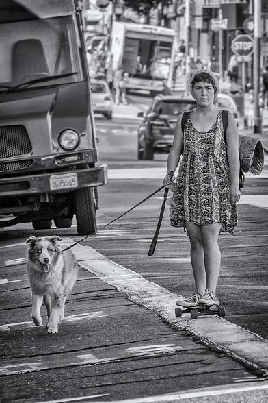 Girl with bedroll on her back skateboards down Haight Street with her dog on a leash.