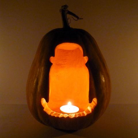 Cousin Itt from The Addams Family, diminutive, floor-length hair being, wearing a bowler hat is depicted on a carved pumpkin. Pumpkin with a tealight candle inside at night.