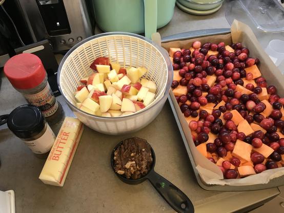 A preparation station featuring diced apples in a colander, a baking tray with a mix of cranberries and sweet potato pieces, and various ingredients including spices, butter, and brown sugar.
