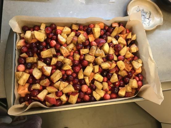 The image features a baking dish filled with a colorful mixture of chopped apples, sweet potatoes, and cranberries, lightly coated with cinnamon & nutmeg. It's lined with parchment paper and placed on a countertop. A small plate is partially visible in the background.