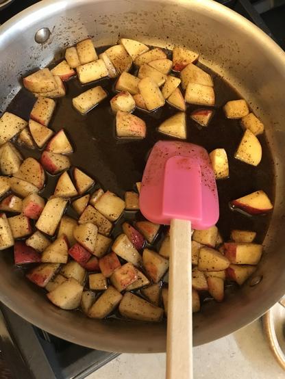A saucepan contains cooked apple chunks in butter, brown sugar & spices, and a pink silicone spatula is resting inside.