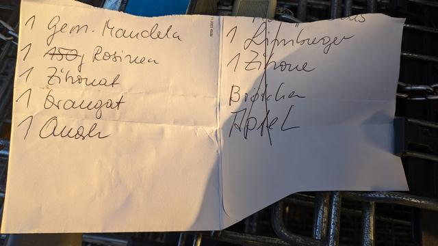 Bildbeschreibung von KI:
Das Foto zeigt eine handgeschriebene Einkaufsliste auf einem Blatt Papier, das auf einem Einkaufswagen liegt. Die Liste ist in zwei Spalten unterteilt:

**Linke Spalte:**
1. Gem. Mandeln (vermutlich gemahlene Mandeln)  
2. ASÖ Rosinen  
3. Zichorie  
4. Braupat  
5. Ausle  

**Rechte Spalte:**
1. Limburger  
2. Zichorie (nochmals aufgeführt)  
3. Brötchen  
4. Apfel (durchgestrichen)  

Das Papier ist leicht geknickt, und im Hintergrund sind Details des Einkaufswagens und dessen Kette sichtbar. Die Liste scheint teilweise abgearbeitet zu sein, da „Apfel“ durchgestrichen wurde.