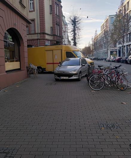 Ein silberner PKW und ein DHL Fahrzeug parken auf einem breiten Gehweg und blockieren ihn komplett.
