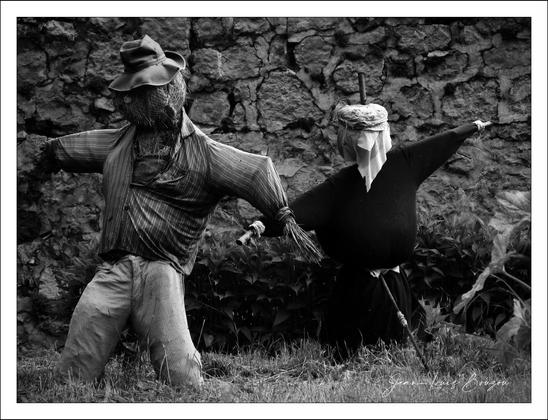 The black and white image shows two scarecrows set against a rough stone wall. The scarecrow on the left is dressed in a striped shirt and pants, with a straw hat on its head. The scarecrow on the right is wearing a dark top and a cloth wrapped around its head with a stick protruding. They are standing amidst some plants and grass, creating a rustic and slightly eerie atmosphere.