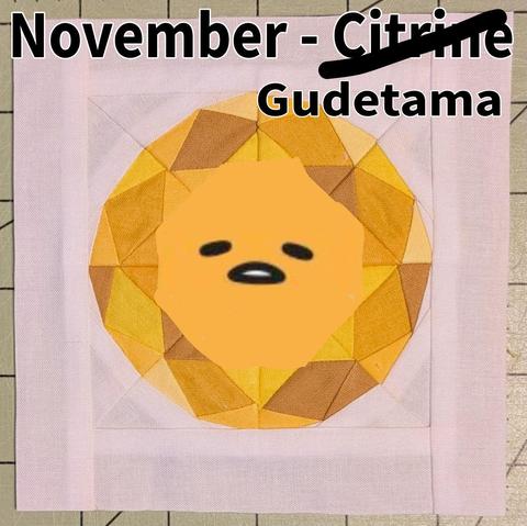 A square quilt block. The sharp edges and angles of the seams give illusion to a faceted gemstone. The gemstone fabric is in shades of light, medium, and dark yellows.   It is surrounded by white fabric to complete the square. It looks vaguely like an egg-- and now Gudetama's lazy face has been drawn in the middle! 
