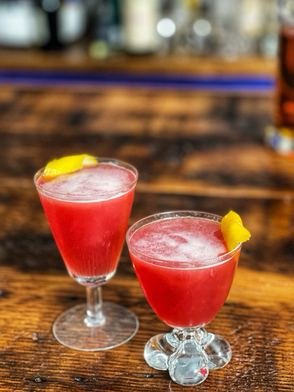 Two cocktails in a coupes sit on a wooden bar top.