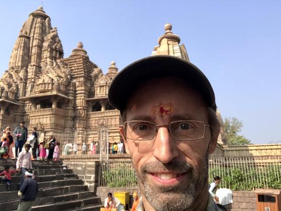 Auf dem Foto bin ich mit Brille und Basecup zu sehen. Auf der Stirn ein roter Punkt sowie gelbes Zeichen. Im Hintergrund ist der Lakshmana Tempel aus dem 10. Jahrhundert zu sehen. Ich bin in Khajuraho.