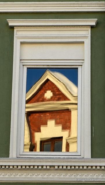 the facade of a red brick and white stucco house is reflected in the window of a green stone house with white window frames and stucco decor below the window