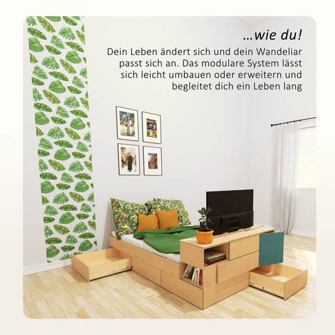 Hier ist ein Bett zu sehen, das am Fußende höher wird, so dass es quasi in eine Art Kommode übergeht. Darauf steht ein Fernseher, der zum Bett gerichtet ist und einige Bücher liegen in den kleineren, offenen Modulen. Die unteren, tiefen Module haben leere Schubladen, die weit geöffnet sind. An der Wand sind vier Bild über dem Kopfende des Bettes sichtbar und die Tapete zeigt einen senkrechten Streifen mit verschiedenen grünen Blättern.